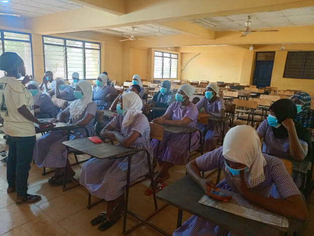 Ein Klassenzimmer in Nigeria mit Schülerinnen und Schülern, die Gesichtsmasken tragen und auf Bänken und dem Boden sitzen, mit Büchern auf Tischen und Fenstern, einer Tür und Deckenventilatoren im Hintergrund.