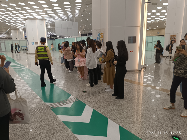 Eine Gruppe von Menschen steht in einer Schlange an einem Flughafen-Sicherheitscheck-in, einige halten Taschen und Mobiltelefone, mit Säulen im Hintergrund und Lampen an der Decke und einem Wasserzeichen am unteren Bildrand.