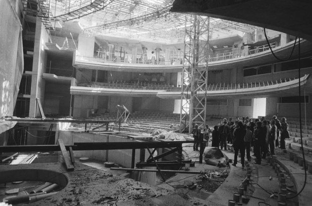 Schwarzes und weißes Bild von Menschen, die auf einem Betonboden in einem unfertigen Gebäude mit Metallpfosten, -rahmen, einer Treppe, einem Geländer, einer Wand und Deckenleuchten stehen.