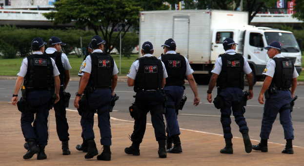 Eine Gruppe von Beamten in Uniform, einschließlich Mützen, Hemden, Schutzwesten, Uhren, Hosen und Schuhen, die auf einer Straße mit einem geparkten Lkw auf der rechten Seite neben einem Zaun, Gebäuden, Bäumen, Pflanzen, Polen und Bannern im Hintergrund und einer Brücke oben rechts gehen.
