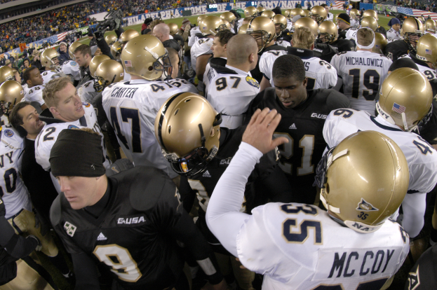 Eine Gruppe von Fußballspielern in Helmen drängt sich auf einem Rasenfeld zusammen, mit einer Menge von Menschen auf den Tribünen und Bannern im Hintergrund.
