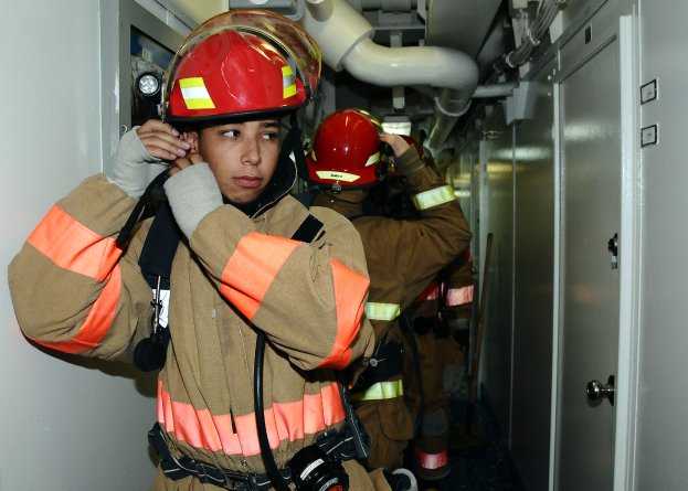Eine Gruppe von Feuerwehrmännern in Uniform, die in einem Raum mit einer Tür auf der rechten Seite und einer Wand auf der linken Seite stehen, mit Rohren und anderen Gegenständen im Hintergrund.