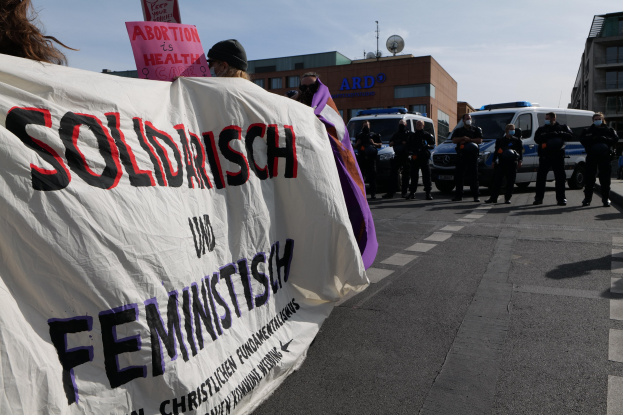 Gruppe von Menschen marschiert auf einer Straße mit einem "Solidarität und Feminismus"-Schild, während parkende Fahrzeuge und Gebäude im Hintergrund zu sehen sind, unter einem klaren blauen Himmel.