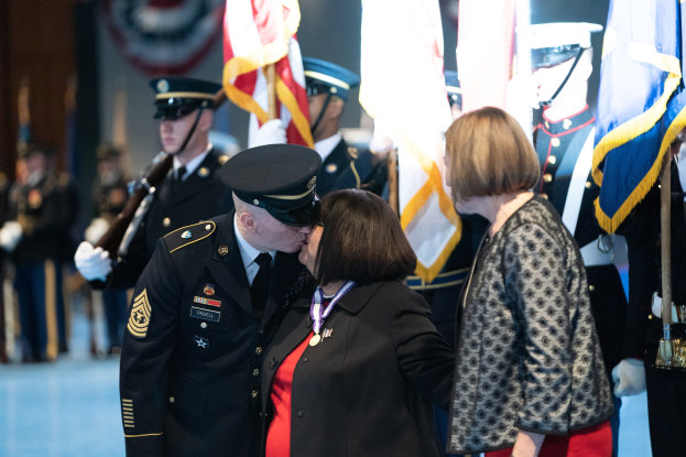 Ein Mann und eine Frau küssen sich, während sie von einer Gruppe uniformierter Personen mit Fahnen umgeben sind.