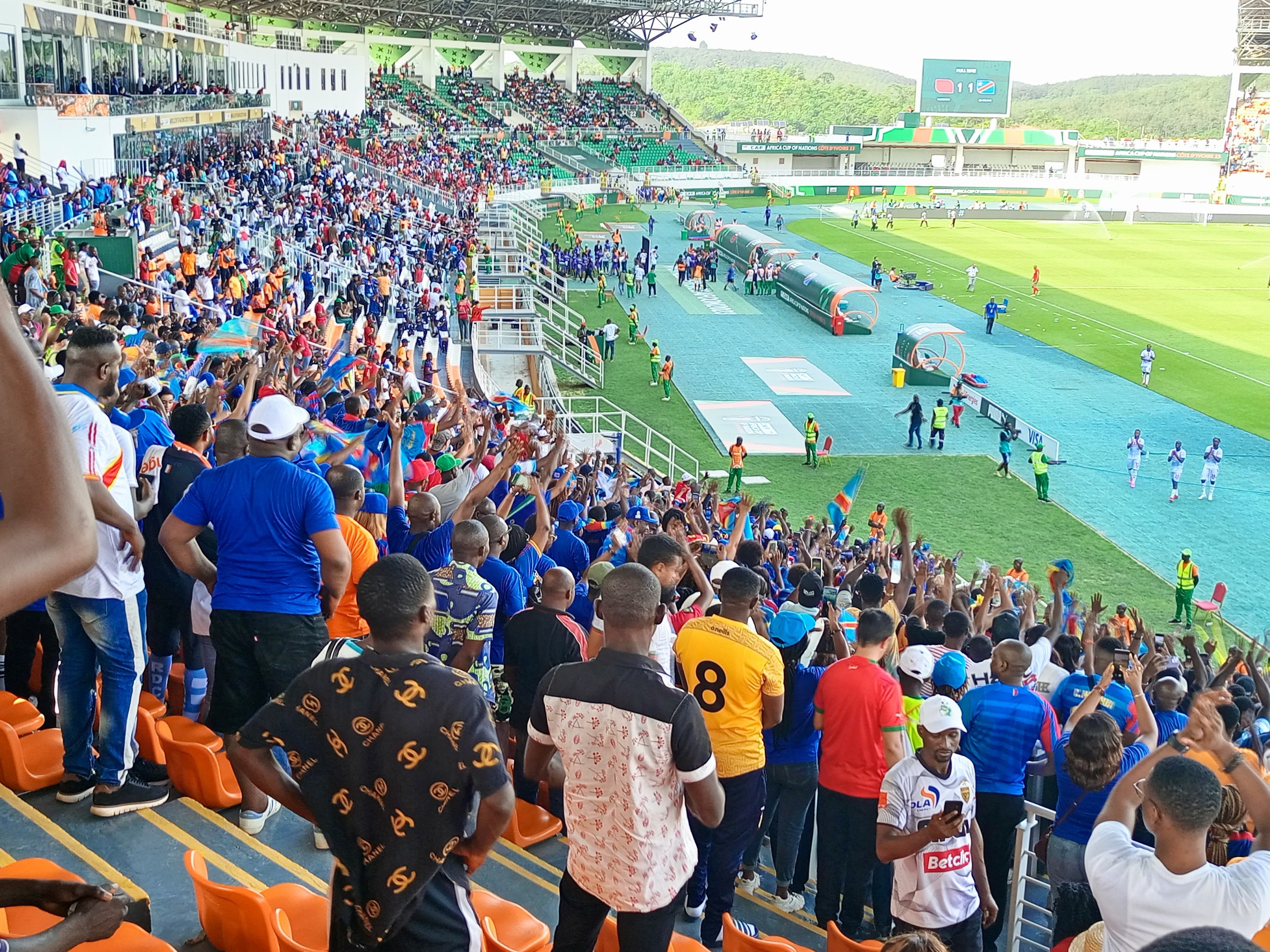 Eine große Menschenmenge versammelt sich vor einem eingezäunten Fußballfeld, wobei einige auf Stühlen sitzen und andere stehen, mit Blick auf Hügel unter einem klaren blauen Himmel während der Afrika-Cup 2019.