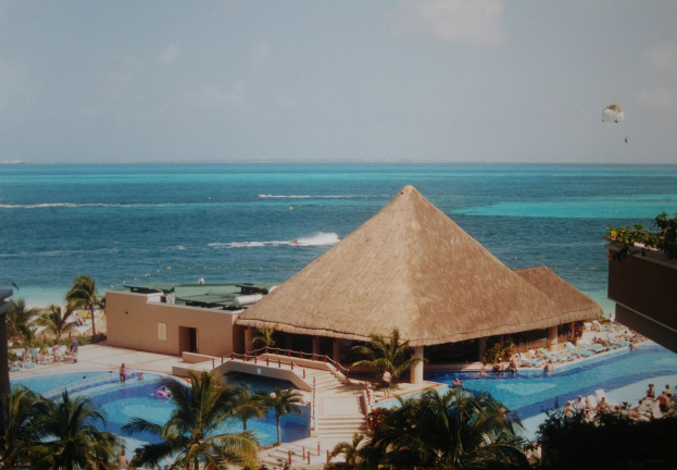 Ferienanlage mit gro├čen Schwimmbad, umgeben von Palmen, Hütten und Menschen, mit einem Ozean und bew├Âlktem Himmel im Hintergrund, mit einem Fallschirm in der Luft.