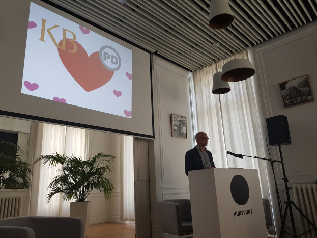 Ein Mann steht an einem Rednerpult mit einem Mikrofon, gibt eine Präsentation vor einer Projektionswand, mit Stühlen, einer Pflanze, Fotorahmen, Vorhängen und Deckenleuchten im Hintergrund.