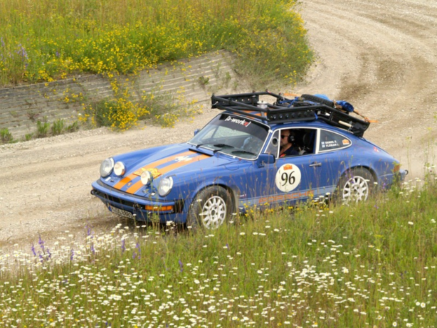 Ein blaues Porsche 911 Carrera RS Rallye-Auto f├Ąhrt auf einer Schotterstra├če durch die Natur, umgeben von Pflanzen und Blumen, mit Text und Zahlen auf der Karosserie und einem sichtbaren Fahrer.