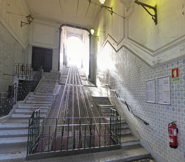 Alte Bahnstation mit Treppe, die zu einer Gebäudeetage führt, mit Geländern, Wandtafeln, Feuerlöscher, Deckenleuchten und einer Tür im Hintergrund.