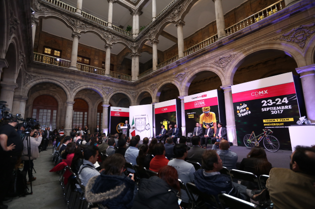 Eine Gruppe von Menschen sitzt auf einem Podium mit Mikrofon vor einer Menge von Menschen, einige halten Kameras. Im Hintergrund gibt es Banner mit Text und Bildern, sowie Säulen, Geländer, Fenster und eine Wand.