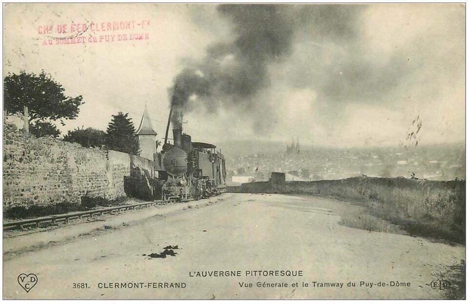 Ein altes Schwarz-Weiß-Foto eines Zuges, der auf Schienen fährt, mit einer Wand auf der linken Seite, Bäumen im Hintergrund und Rauch, der aus der Lokomotive aufsteigt, mit Text oben und unten, der 'L'auvergne pittoresque - vue générale et le tramway du puy-de-dôme' lautet.