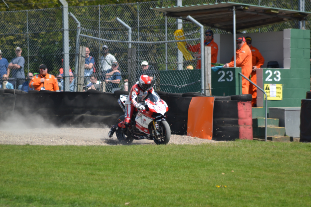 Eine Person auf einem Motorrad fährt auf einem grasbewachsenen Feld, mit Zuschauern in Mützen und Schutzbrillen, einem Maschendrahtzaun, Pfählen, Bäumen, Treppen mit Geländern und einem Schuppen mit Zahlen und Texttafeln im Hintergrund.