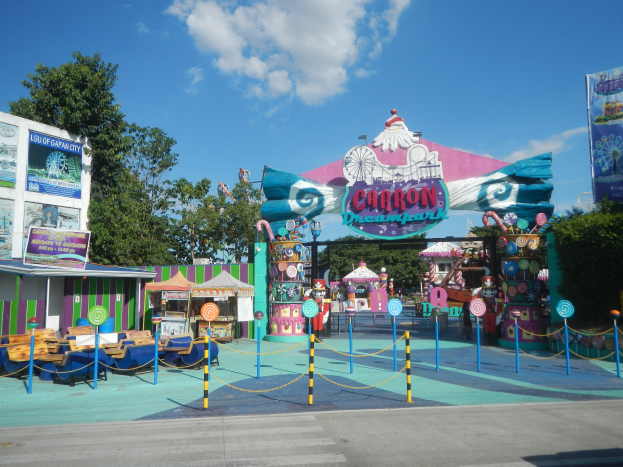 Karussell am Parkeingang mit Straße darunter, eingezäuntem Bereich, Buden, Bannern, Bäumen und bewölktem Himmel.