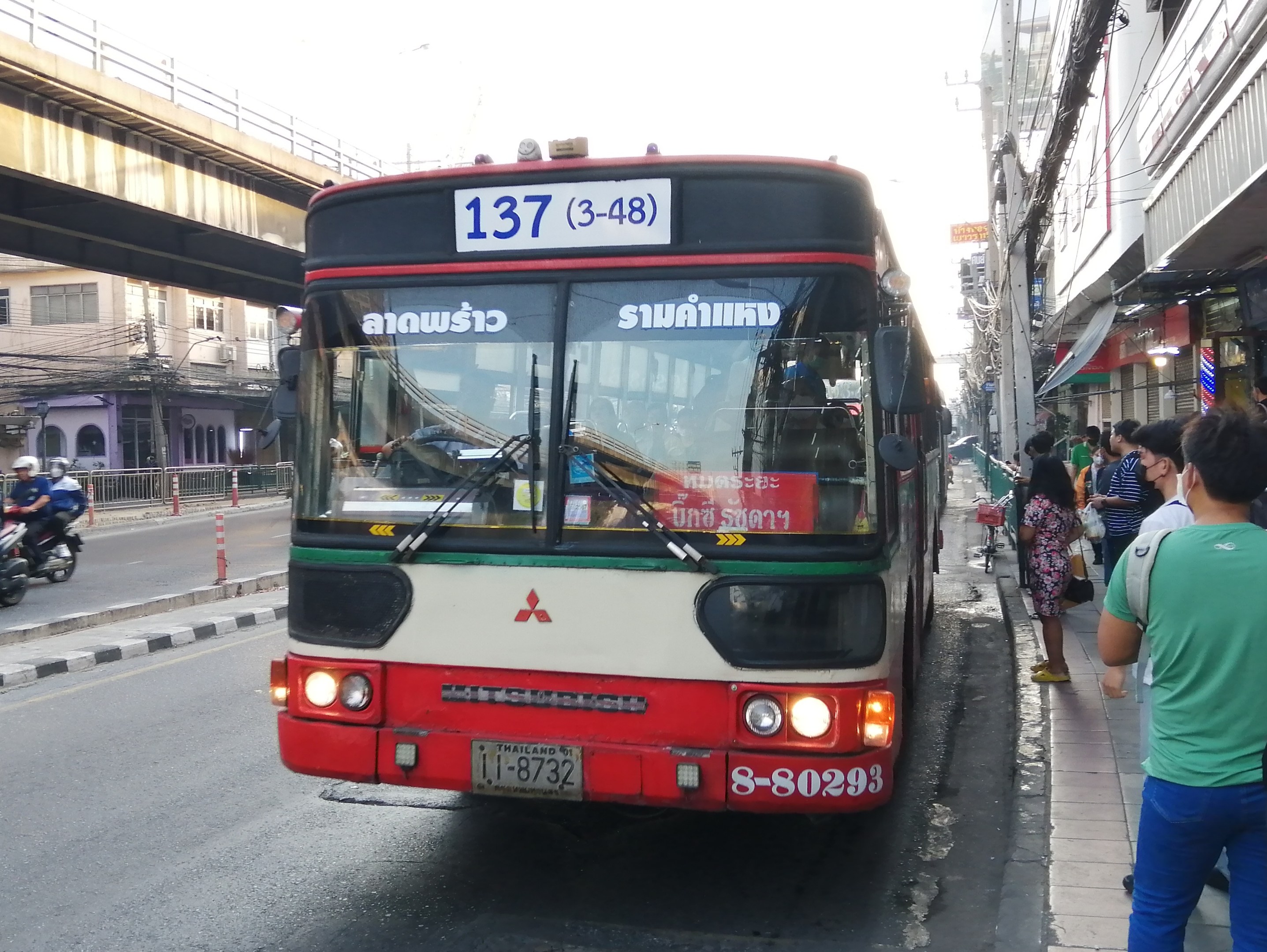 Ein roter und weißer Bus fährt auf einer Straße mit hohen Gebäuden, mit Passanten auf dem Gehweg und einer Brücke im Hintergrund unter einem klaren blauen Himmel.