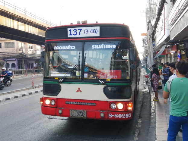 Ein roter und weißer Bus fährt auf einer Straße mit hohen Gebäuden, mit Passanten auf dem Gehweg und einer Brücke im Hintergrund unter einem klaren blauen Himmel.