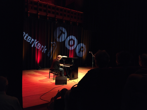 Ein Mann mit Mütze sitzt an einem Piano auf einer schwach beleuchteten Bühne, rechts daneben ein Mikrofonständer und ein Vorhang mit Text im Hintergrund.