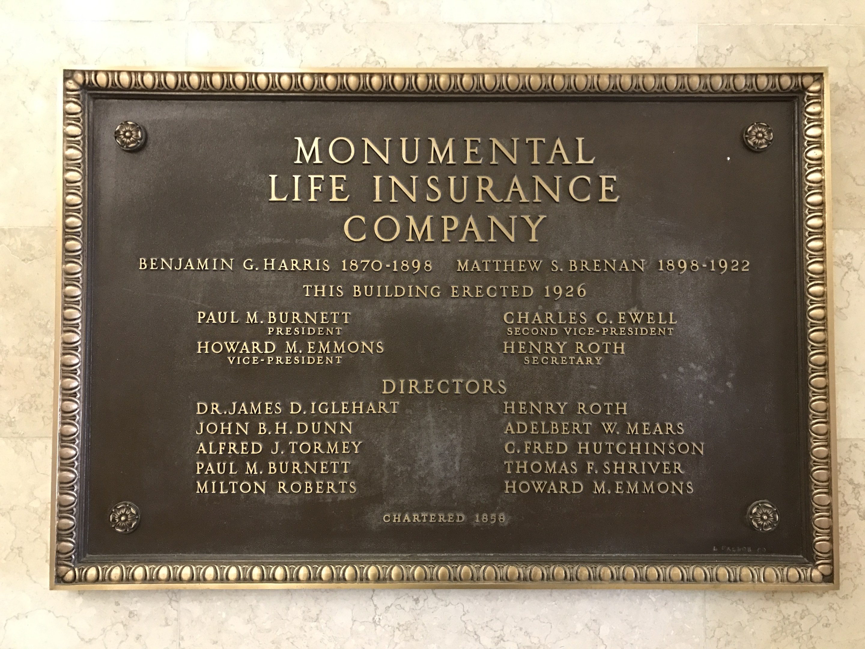 Plakette an einer Gebäudewand mit der Aufschrift "Monumentale Lebensversicherungsgesellschaft" in fetter Schrift, umgeben von einem dekorativen Rahmen, auf einem dunklen Material montiert.