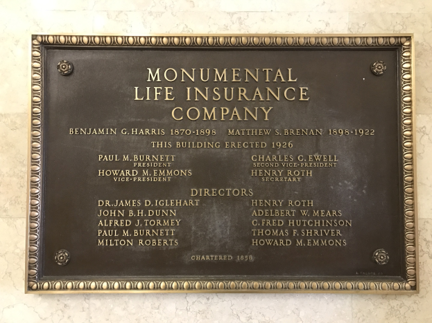 Plakette an einer Gebäudewand mit der Aufschrift "Monumentale Lebensversicherungsgesellschaft" in fetter Schrift, umgeben von einem dekorativen Rahmen, auf einem dunklen Material montiert.