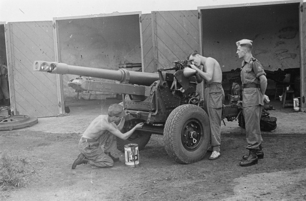 Zwei Männer in Uniformen und Mützen arbeiten an einer großen Artilleriekanone vor einem Gebäude, mit einer Hütte und anderen Gegenständen im Hintergrund, schwarz-weiß.