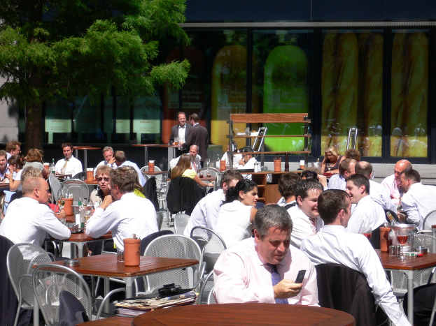 Eine Kantine mit Gruppen von Menschen an Tischen sitzend und sich unterhaltend, zwei Individuals in schwarzen Anzügen in der Mitte stehend, ein Flaschencontainer rechts, ein Baum links und Saftgläser und Flaschen auf den Tischen.