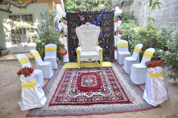 Eine Gartenhochzeitsaufstellung mit weißen Stühlen, rotem Teppich, Blumenbouquets, Dekorationsgegenständen, Ballons, Pflanzen, Bäumen, einem Gebäude mit Fenstern und Tür und einer Wand auf der rechten Seite.