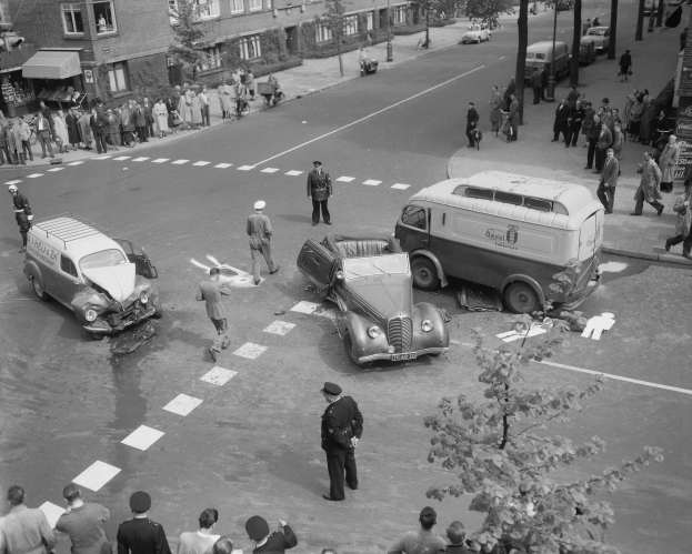 Schwarz-weißes Bild einer Autounfall-Szene an einer Straßenkreuzung mit mehreren Fahrzeugen, darunter ein Lieferwagen, und einer Gruppe von Schaulustigen; Gebäude, Bäume, Pfähle und Schilder sind im Hintergrund zu sehen.