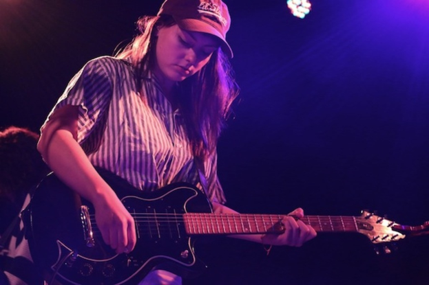 Eine Frau mit Mütze spielt Gitarre auf der Bühne eines Musikkonzerts, während zahlreiche Lichter den Hintergrund erhellen.