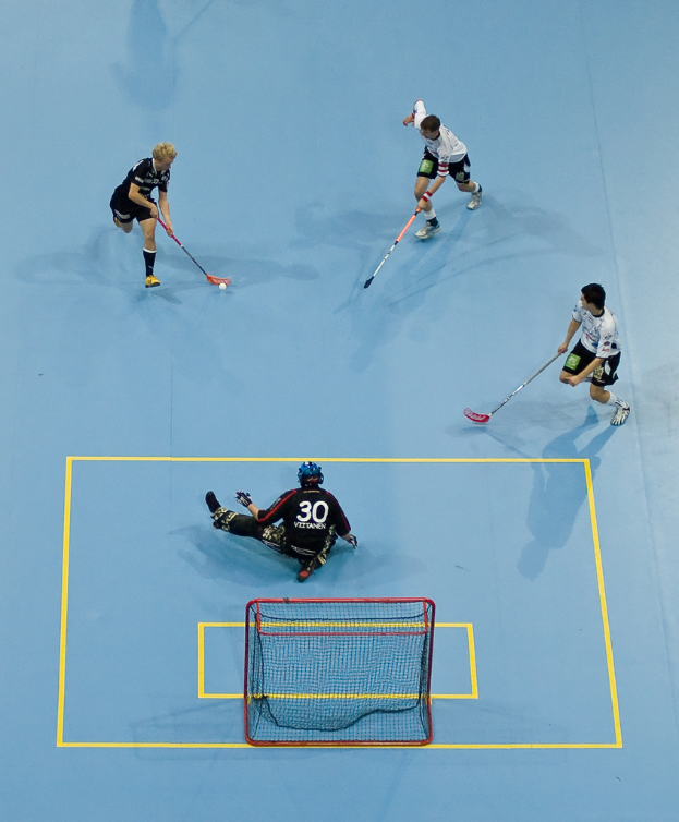 Drei Personen in Sportkleidung spielen Hockey, wobei eine Person auf der rechten Seite den Ball mit einem Hockeystock schlägt und eine Person hinter einem Netz auf dem Boden sitzt.