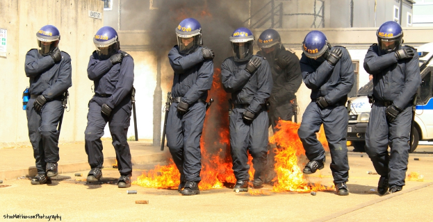 Menschen in Helmen stehen vor einem Feuer, mit Gebäuden, einem Fahrzeug und diversen Gegenständen im Hintergrund; ein Plakat und eine Tafel sind an der linken Wand, und Text befindet sich am unteren Rand des Bildes.