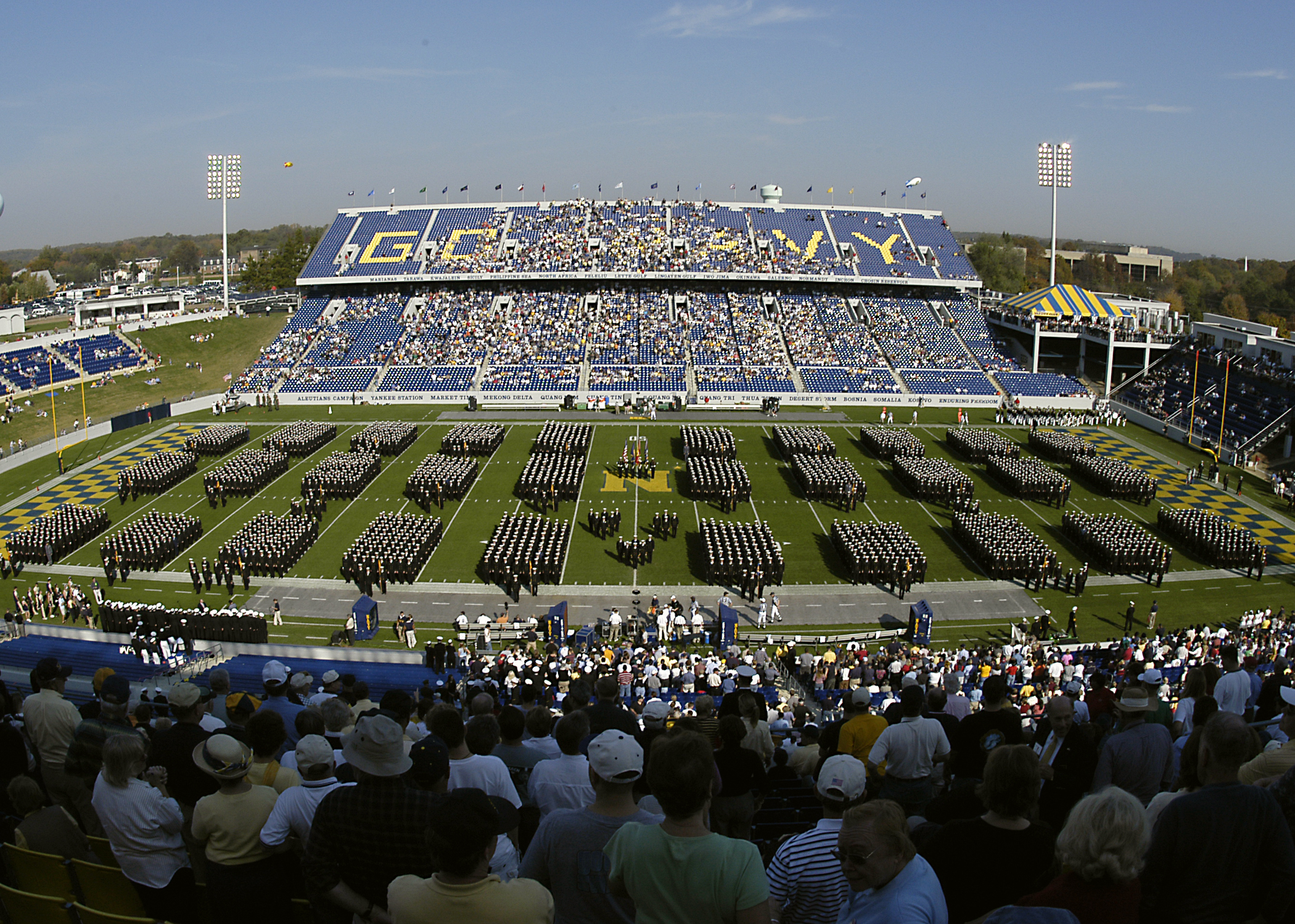 Eine große Menschenmenge in einem Stadion bei einem Fußballspiel, wobei einige stehen und andere sitzen, auf einem grasbewachsenen Feld unter einem klaren blauen Himmel mit Laternen, Bäumen und Gebäuden im Hintergrund.