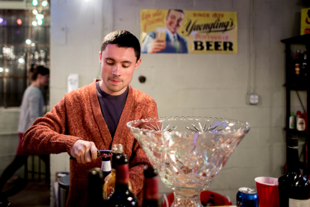 Ein Mann versucht, eine Flaschenkappe mit einem Öffner hinter sich zu öffnen, ein großes Glasregal, ein Regal mit Flaschen und ein Plakat an der Wand im Hintergrund.
