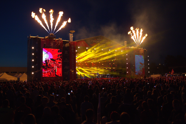 Eine große Menschenmenge auf einem Musikfestival mit einer beleuchteten Bühne mit Performern, umgeben von Bannern, Zelten links und Feuerwerk in einem dunklen Himmel.