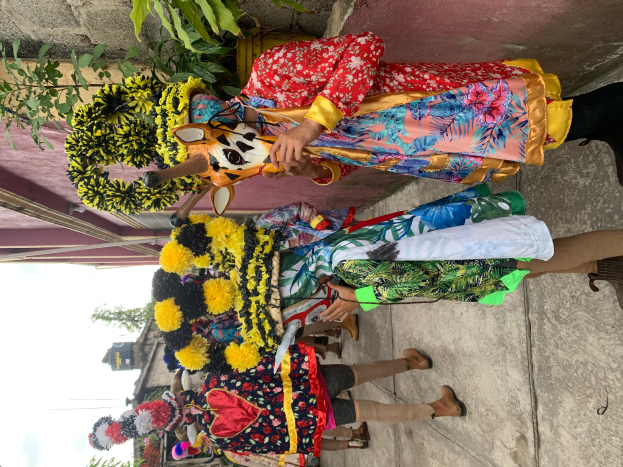 Eine Gruppe von Menschen in farbenfrohen Kostümen steht zusammen vor einem Gebäude, mit verschiedenen Gegenständen, Bäumen, Gebäuden und einem klaren blauen Himmel im Hintergrund bei einem Karnevalsumzug.