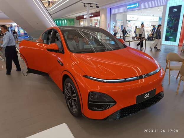 Orangefarbener Elektroauto in einer Ausstellungsfläche umgeben von Menschen, mit Stühlen und einem Wagen rechts und Geschäften im Hintergrund.