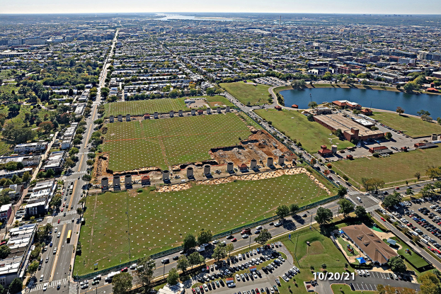 Eine Luftaufnahme einer Stadt mit Gebäuden, Bäumen, Straßen mit Fahrzeugen, Wasser und einem bewölkten Himmel, die einen großen zentralen Platz für einen Park vorschlägt.