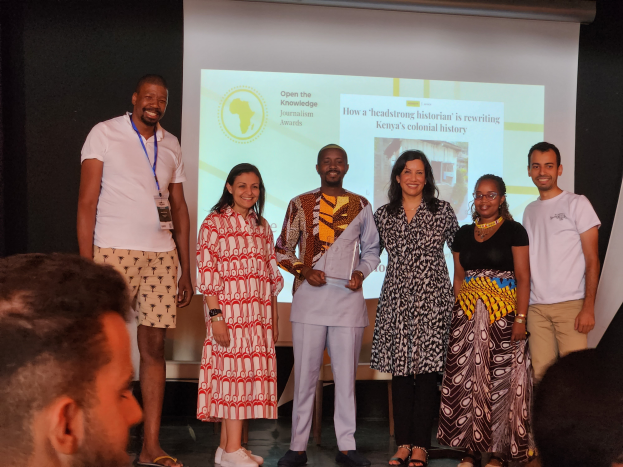 Eine Gruppe von Menschen auf einer Bühne, die lächeln und für ein Foto posieren, mit einem Publikum vor ihnen und einem Bildschirm im Hintergrund, der 'Open the Knowledge Journalism Awards 2019' anzeigt.