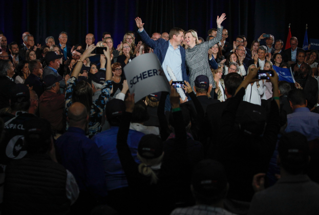 Ein Mann und eine Frau stehen vor einer Menge mit Schildern und Handys, mit einem Vorhang und Fahnen dahinter auf einer Wahlveranstaltung.