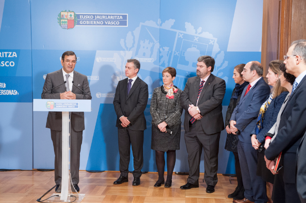Ein Mann im Anzug und mit Krawatte steht hinter einem Podium mit sichtbarer Abnutzung, flankiert von einer weiteren Person im schwarzen Anzug und einer Frau, sowie einigen Personen im Hintergrund und einem Banner dahinter.